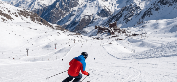 Person skiing down snowy mountain slope with ski resort and snowy peaks in the background